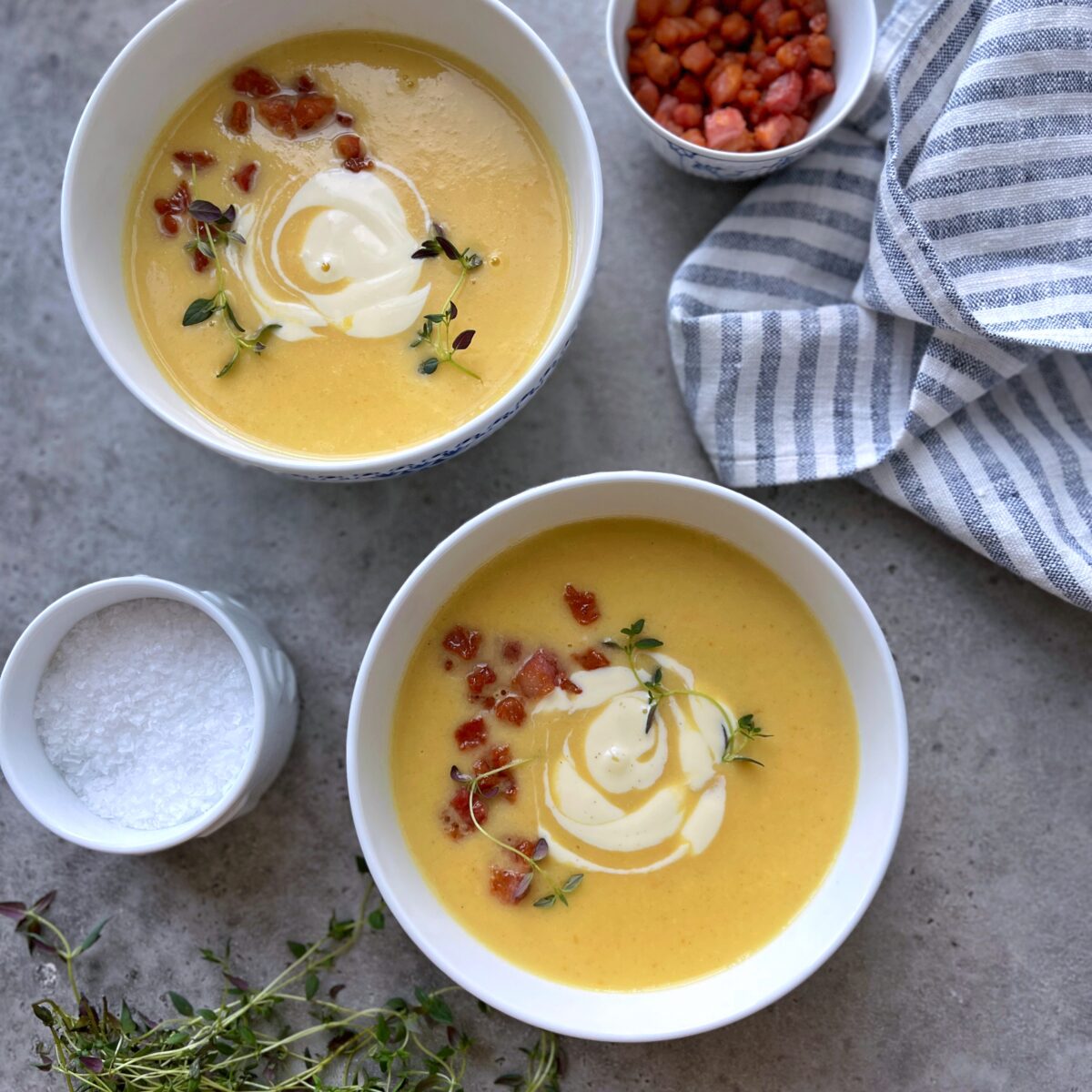 Two bowls of creamy vegetable soup topped with crème fraîche, crispy bacon bits, and thyme, with a bowl of salt, a cup of diced bacon, and a striped cloth nearby on a gray surface.