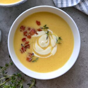 A bowl of creamy yellow veggie soup topped with crispy bacon bits, a swirl of sour cream, and fresh thyme sprigs, placed on a gray surface next to a striped napkin.