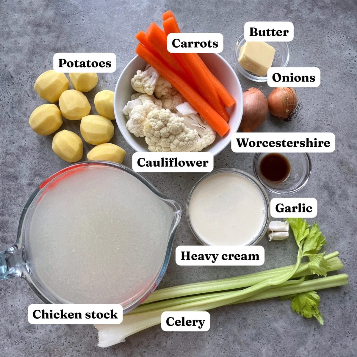 A variety of soup ingredients including peeled potatoes, carrots, cauliflower, butter, onions, Worcestershire sauce, garlic, heavy cream, chicken stock, and celery.