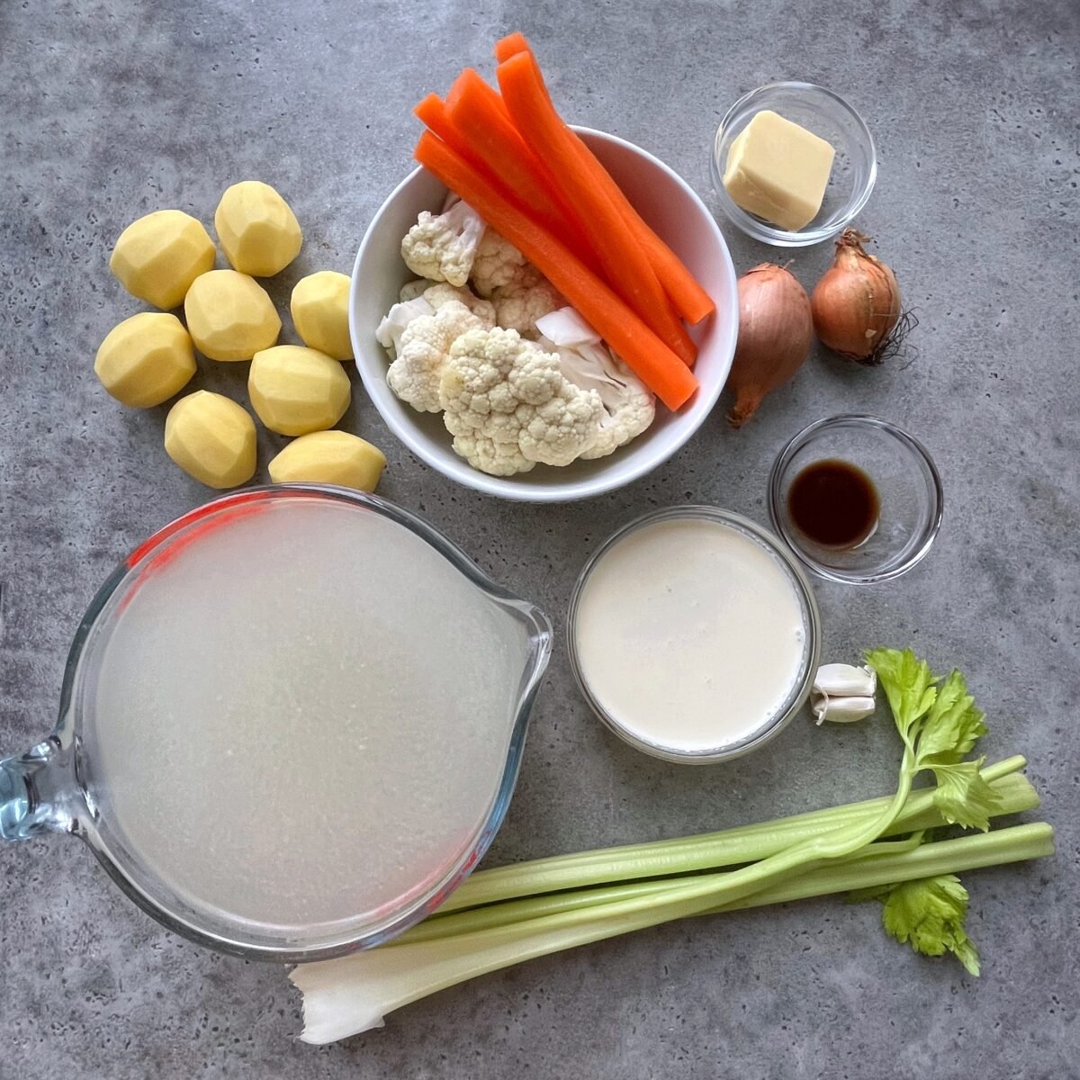 Ingredients for a vegetable soup arranged on a gray surface, including peeled potatoes, carrots, cauliflower, celery, cream, broth, butter, onions, and garlic.