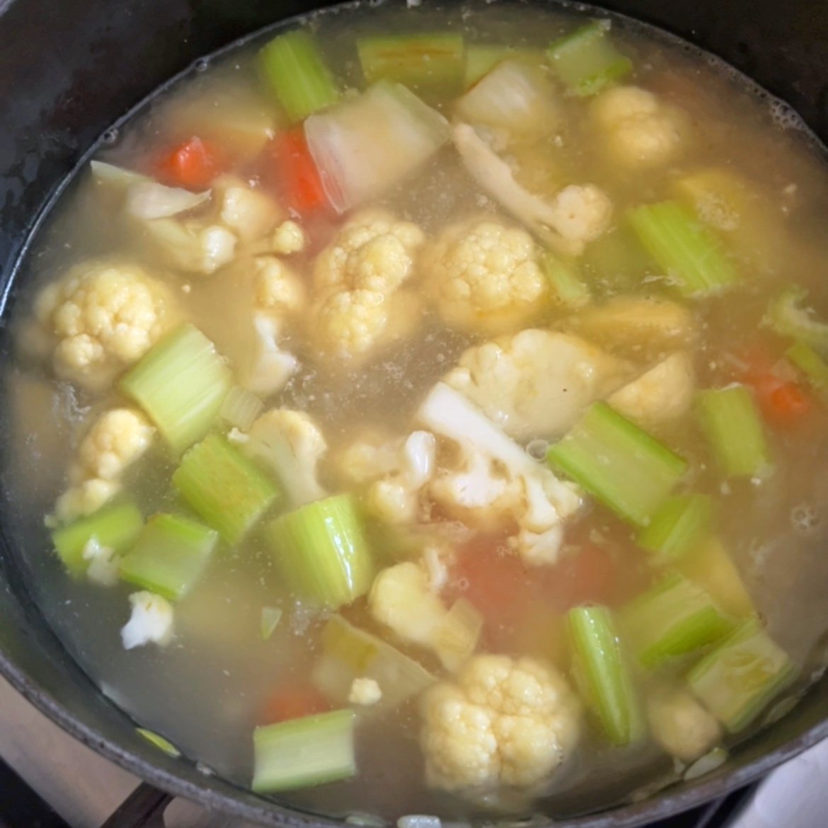 A pot of creamy vegetable soup with visible cauliflower, celery, carrots, and stock simmers gently on the stovetop.