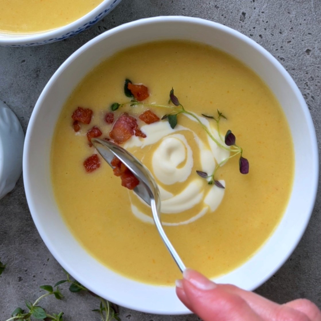 A hand holds a spoon over a bowl of creamy vegetable soup, its yellow surface garnished with diced bacon, a swirl of heavy cream, and fresh thyme.