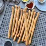 Breadsticks topped with sesame seeds are arranged on a cooling rack, with small bowls of olive oil, vinegar, and seeds nearby on a grey surface.