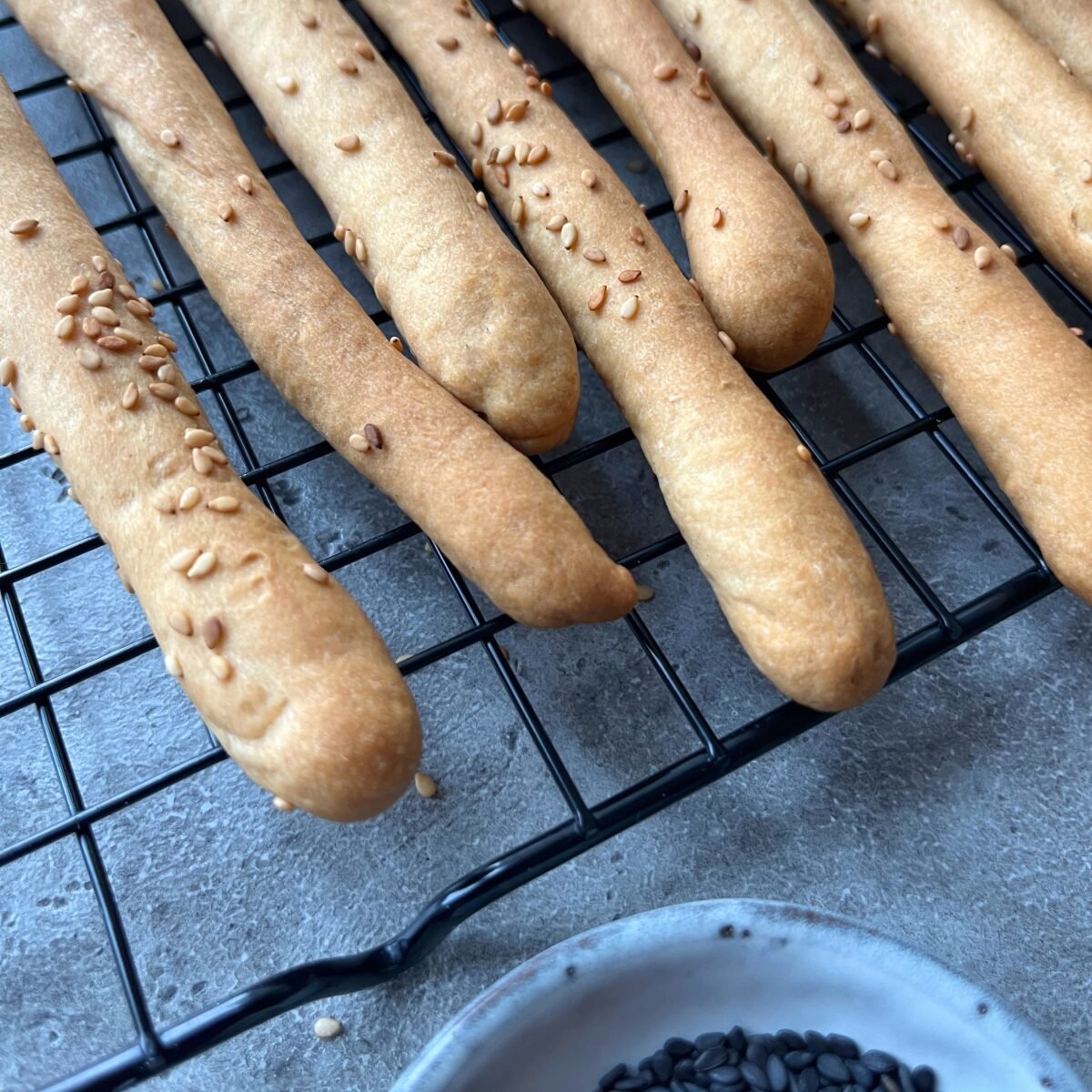 Several golden-brown breadsticks topped with sesame seeds are cooling on a wire rack, with a small dish of black sesame seeds nearby.