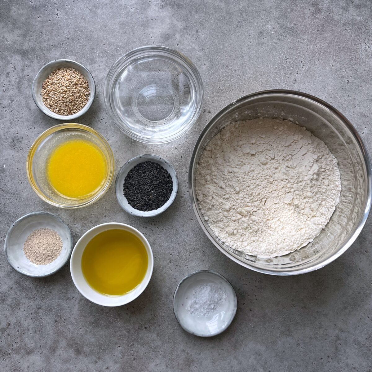 Various ingredients for breadsticks, including flour in a bowl, melted butter, olive oil, yeast, sesame seeds, black seeds, baking powder, and a bowl of water, arranged on a gray surface.