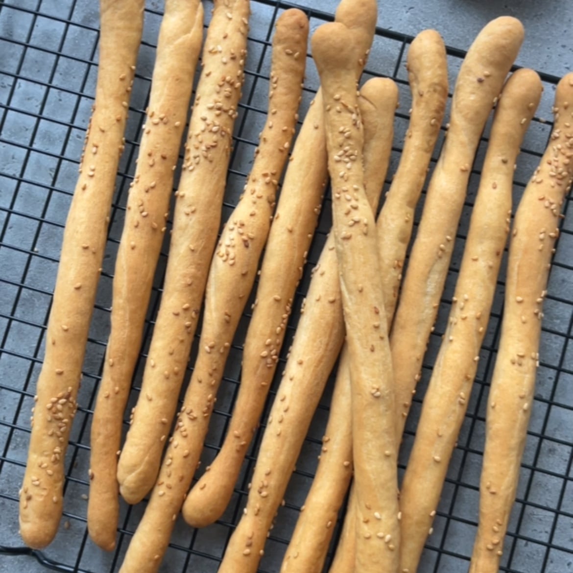 Several golden-brown homemade breadsticks, some topped with sesame seeds, are arranged on a black cooling rack.
