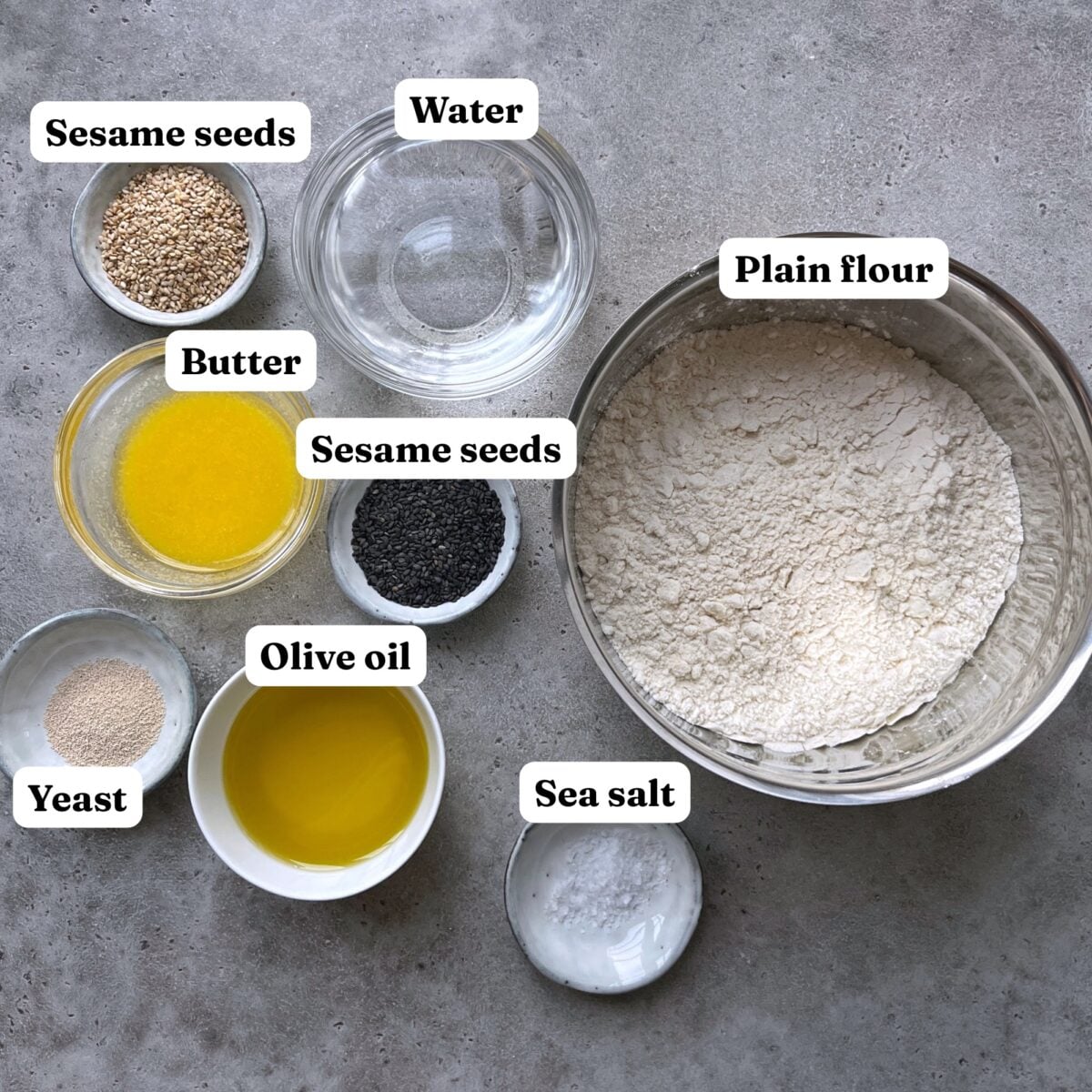 A bowl of flour, small bowls of water, sea salt, olive oil, yeast, melted butter, and two bowls of sesame seeds (white and black) are arranged on a gray surface.