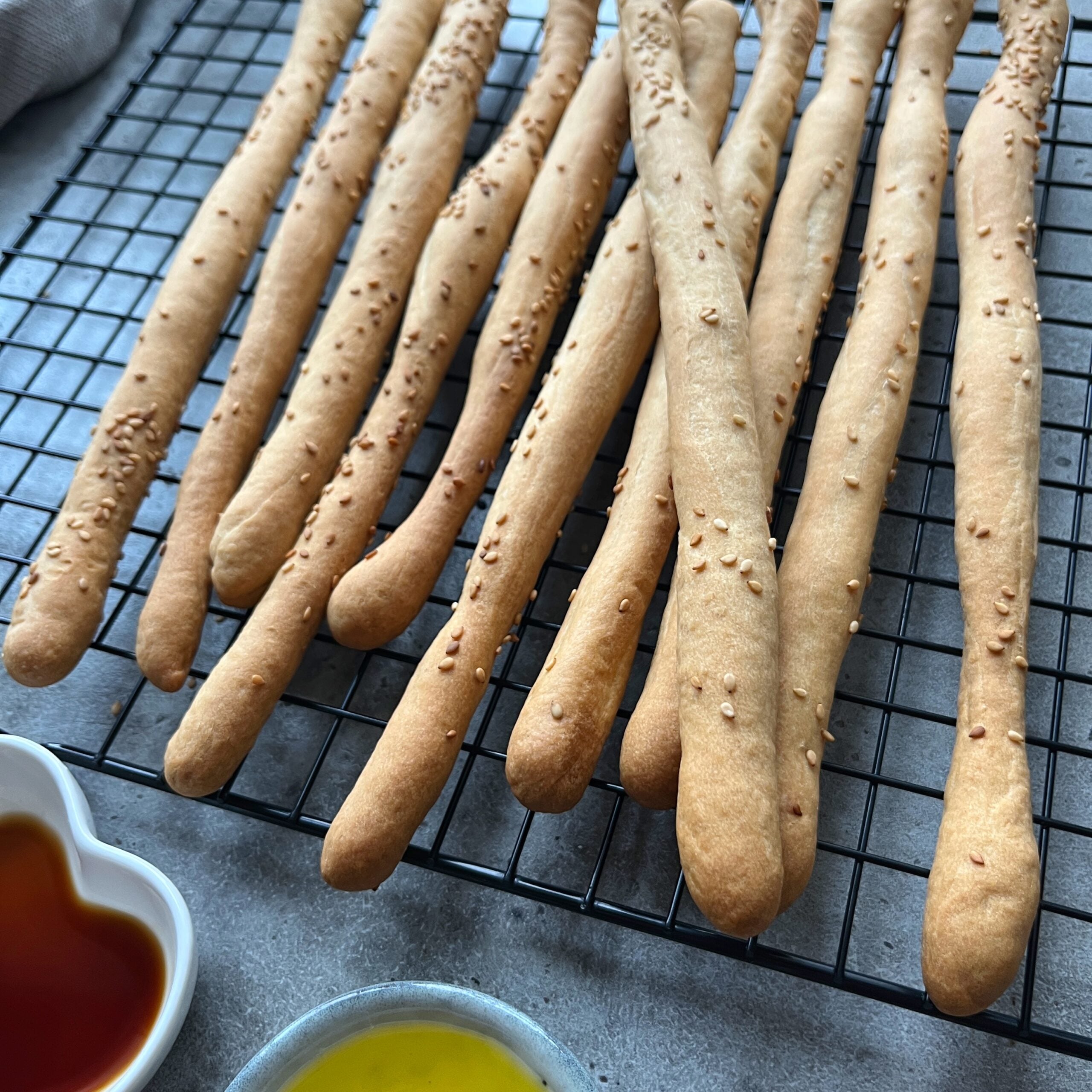 A batch of homemade breadsticks topped with sesame seeds cools on a black wire rack, with small bowls of olive oil and red dipping sauce nearby.
