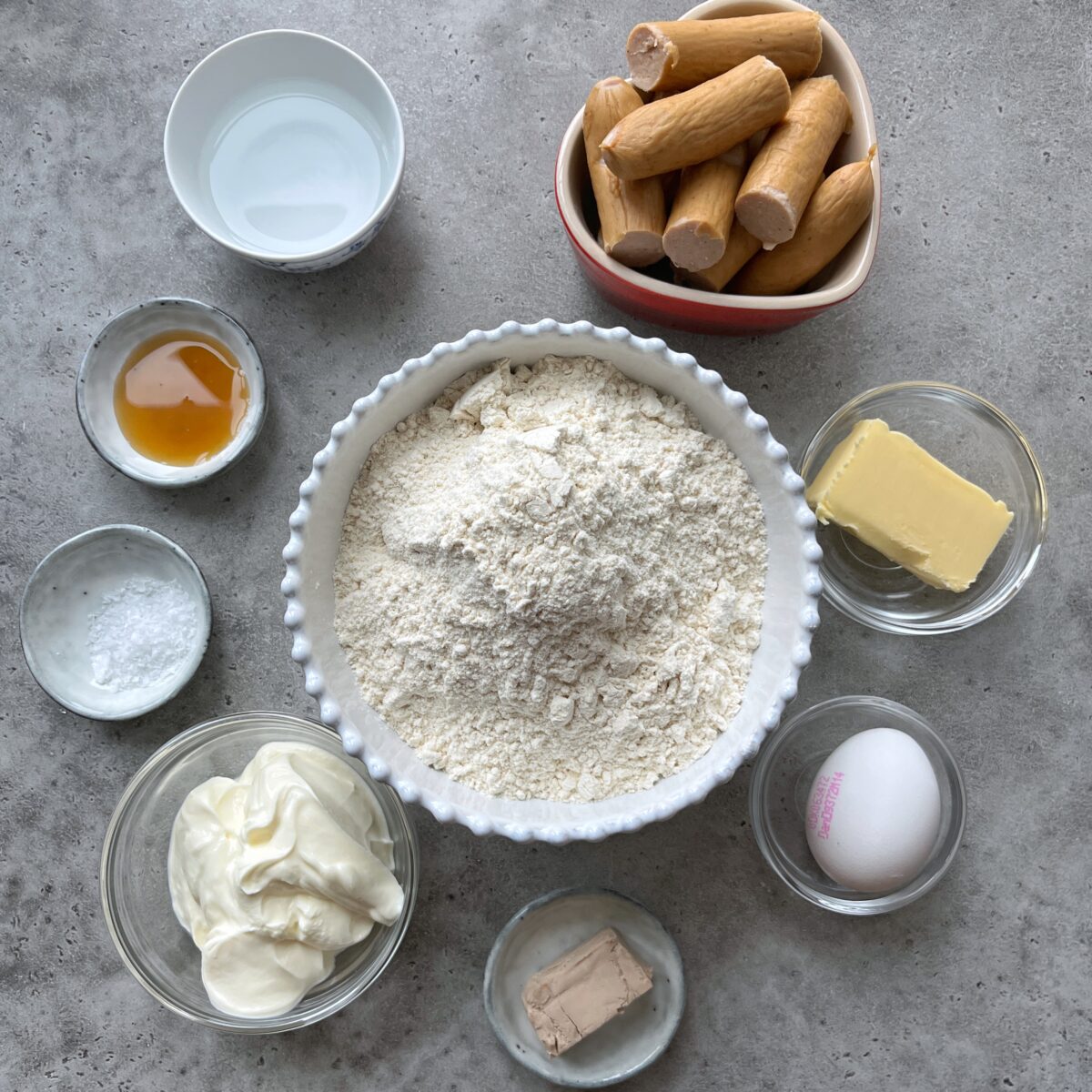 Assorted baking ingredients in bowls on a grey surface, including flour, butter, maple syrup, sour cream, baking powder, yeast, water, egg, and several uncooked sausages.