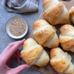 A hand reaches for a golden, sesame-topped homemade sausage roll on a wooden board, with more rolls nearby and a small bowl of sesame seeds on a gray surface.