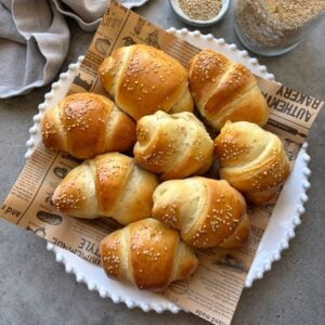 A plate featuring baked sausage rolls on printed parchment paper. In the background is a jar of sesame seeds and a grey kitchen towel.
