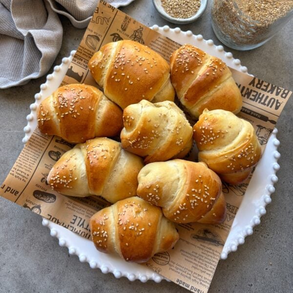 A plate featuring baked sausage rolls on printed parchment paper. In the background is a jar of sesame seeds and a grey kitchen towel.