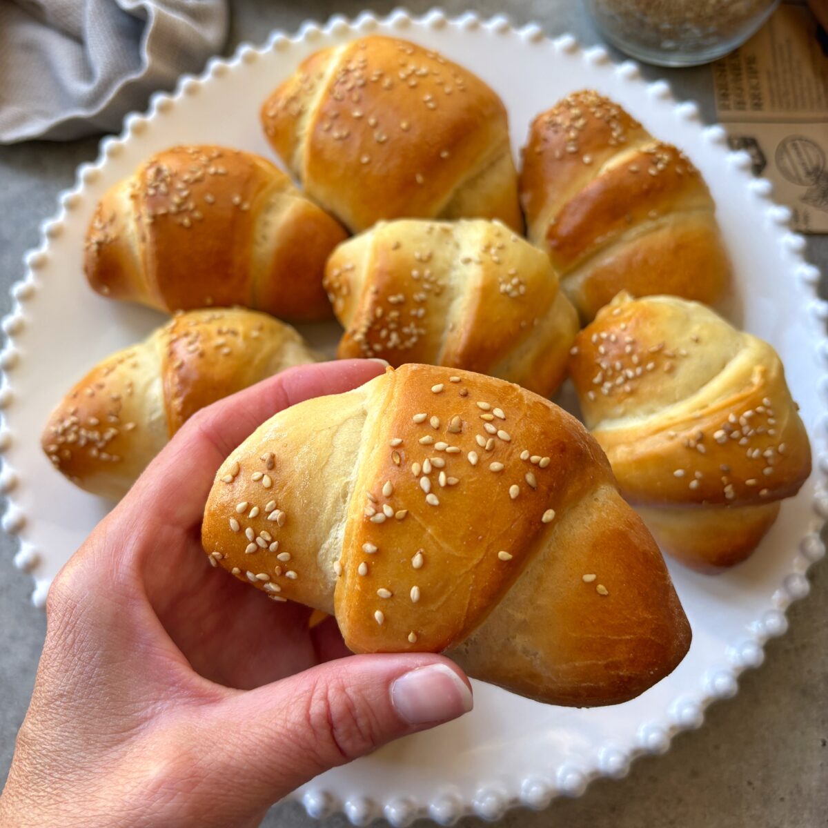 A hand holds a golden brown sausage rolls. In the background is a fluted plate of more rolls.