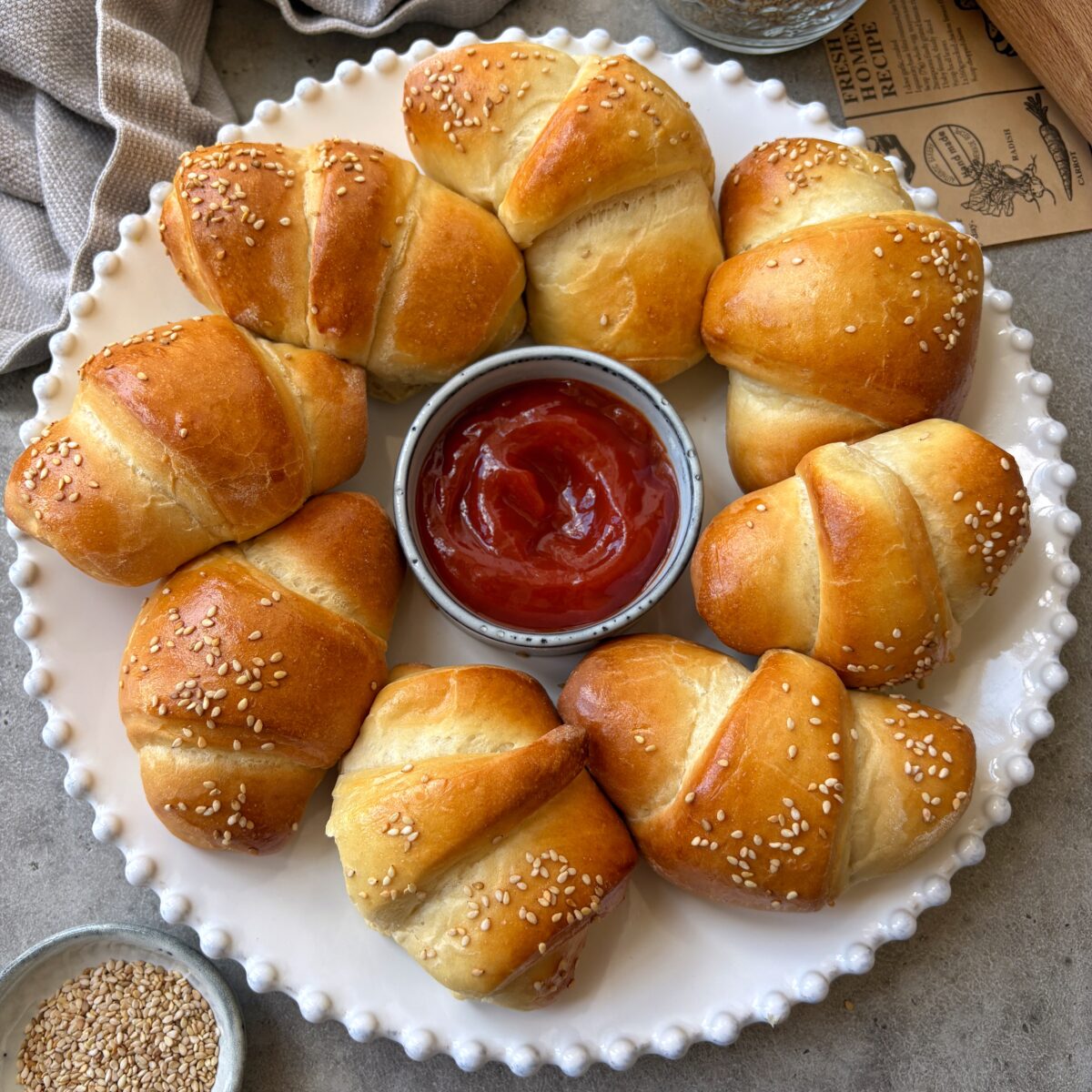 A plate of golden brown sausage rolls topped with white sesame seeds, with a light blue bowl of ketchup in the center.