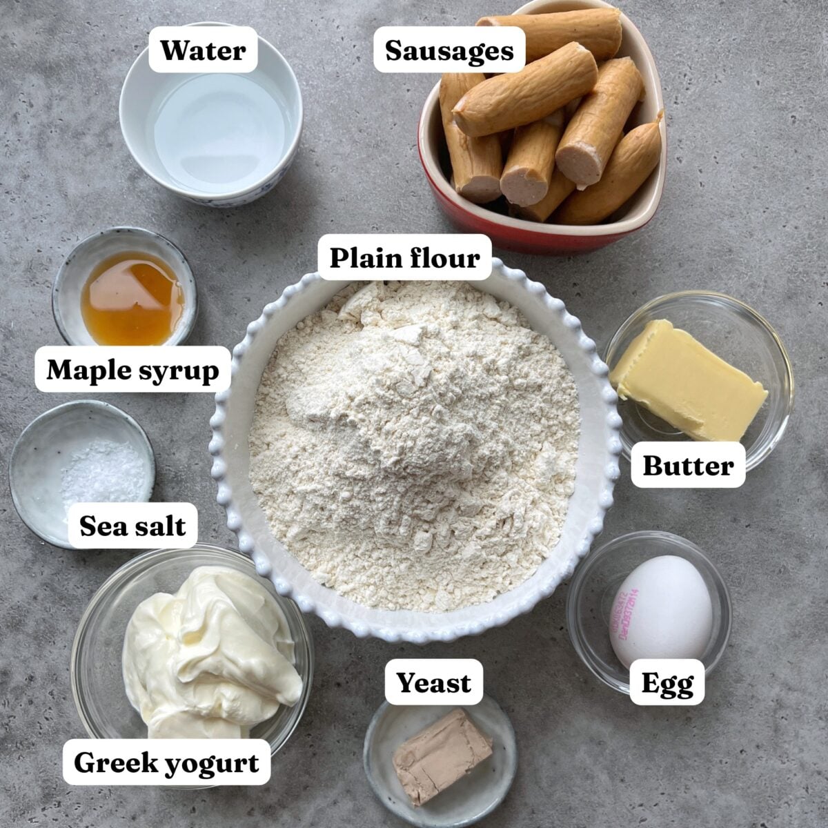 Bowl of flour surrounded by labeled ingredients including water, sausages, maple syrup, butter, egg, yeast, Greek yogurt, and sea salt on a gray surface.