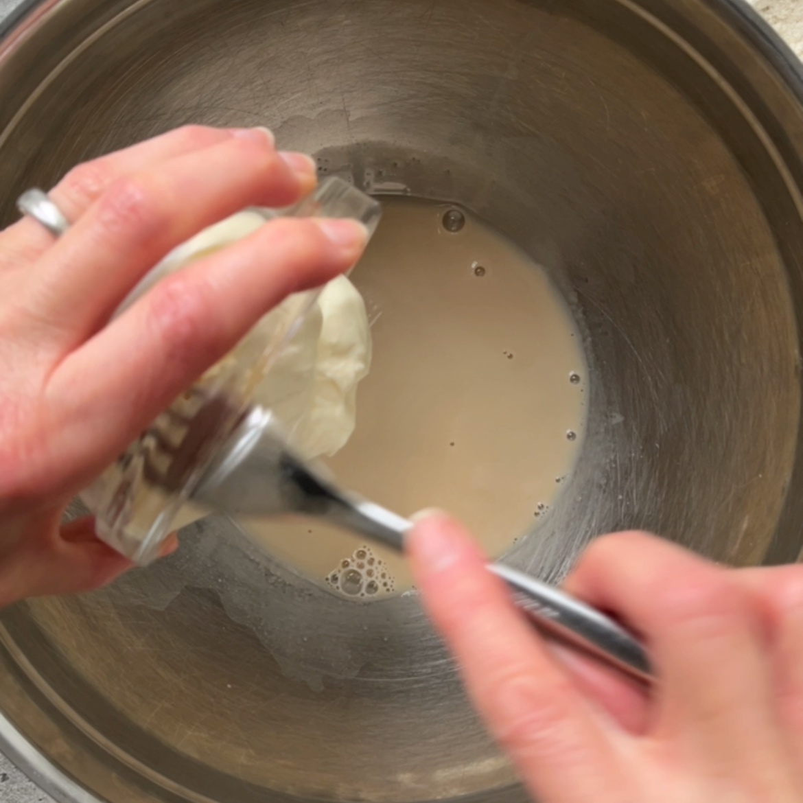 Hands adding a Greek yogurt from a small bowl into a metal mixing bowl containing water and yeast.