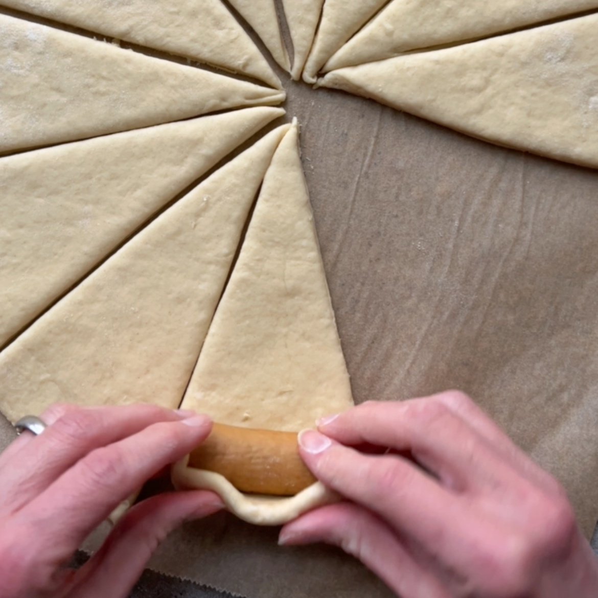Hands rolling a sausage into a triangular piece of dough from the wide end, with several other dough triangles arranged in a circle on parchment paper.