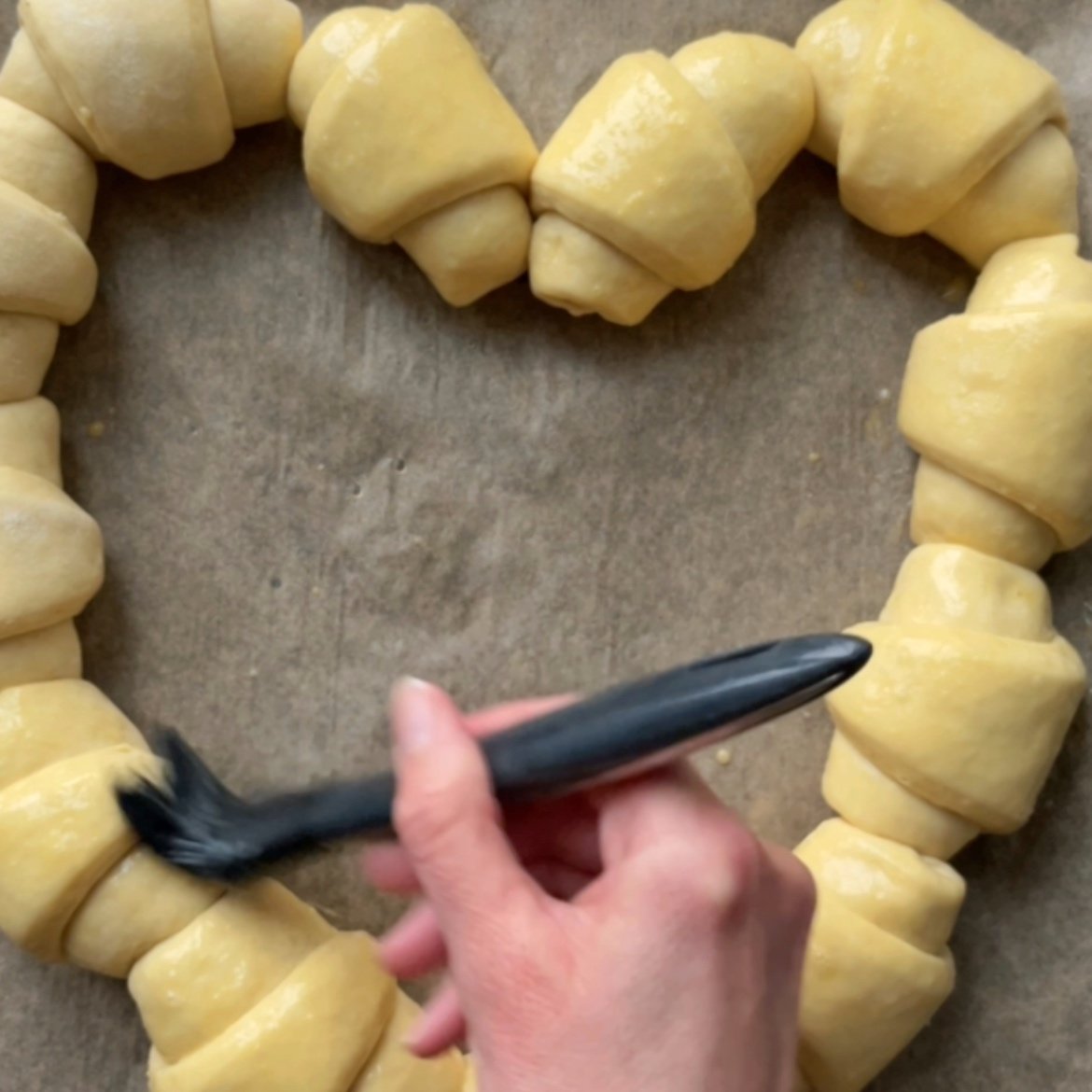 A hand uses a brush to apply egg wash to unbaked sausage rolls arranged in a heart shape on parchment paper.