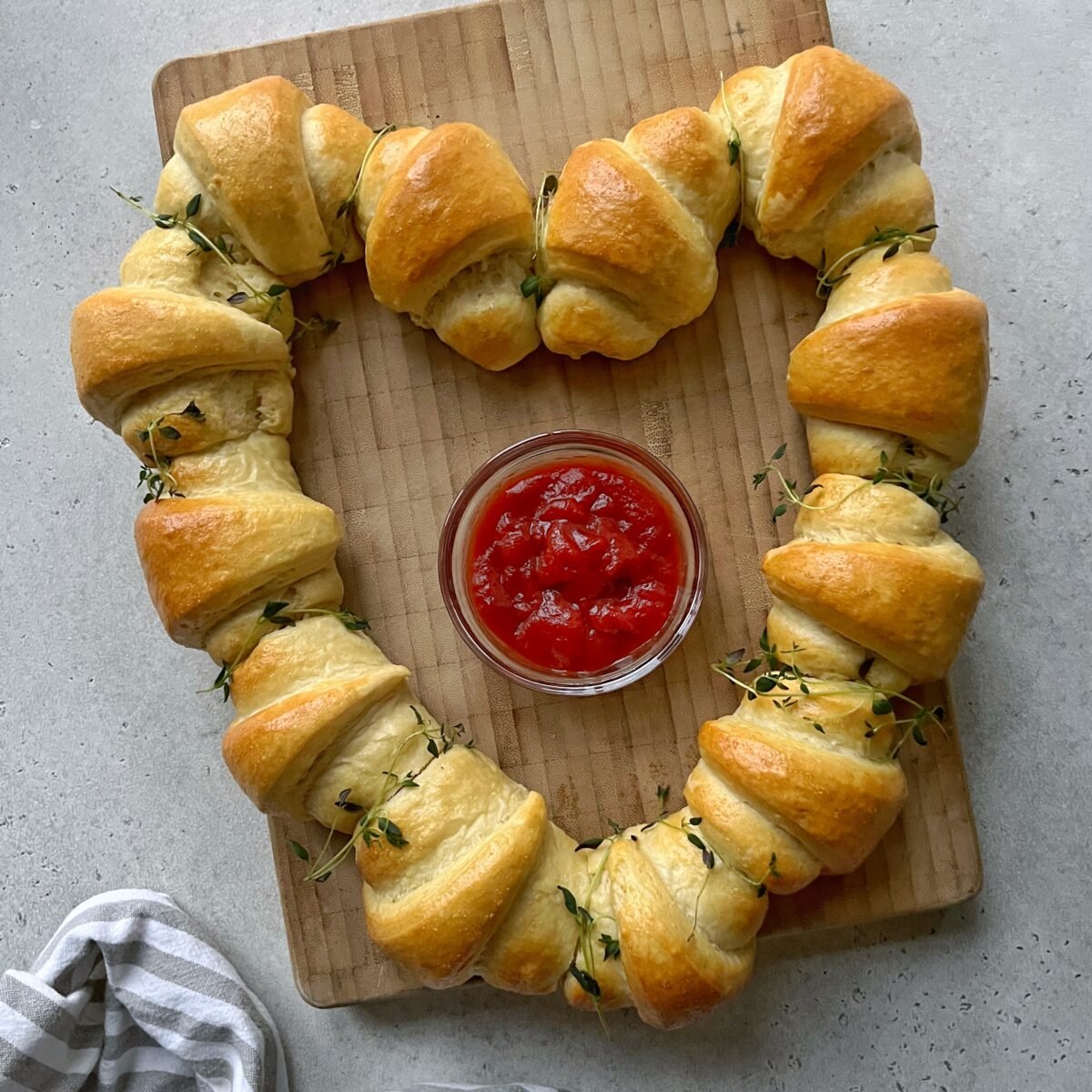 A heart-shaped ring of baked sausage rolls on a wooden board surrounds a bowl of ketchup.