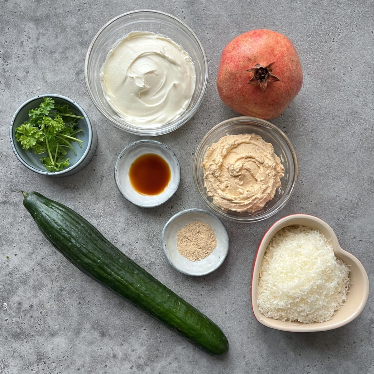 A flat lay of cucumber, parsley, pomegranate, two types of cream cheese, shredded Parmesan, Worcestershire sauce, and a small bowl of garlic powder on a gray surface.