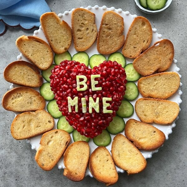 A plate with toasted bread slices arranged around cucumber slices and a heart-shaped cheese ball topped with pomegranate seeds and the words BE MINE spelled with cucumber letters.