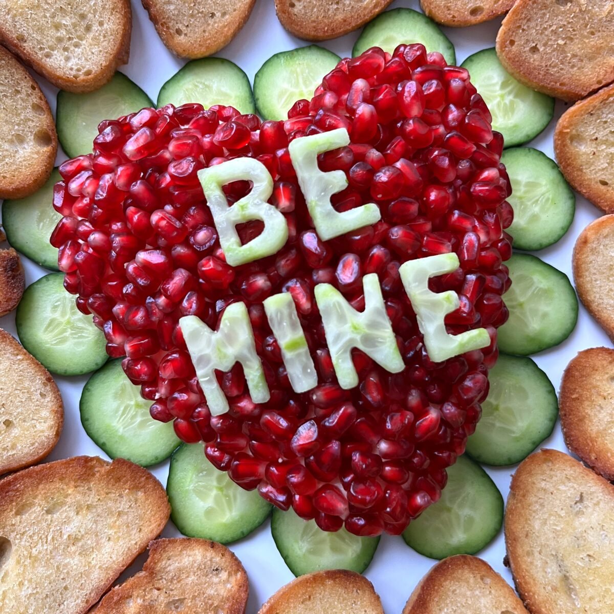 Heart-shaped cheese ball arrangement with pomegranate seeds and the words BE MINE spelled in cucumber slices, surrounded by cucumber rounds and toasted bread slices.