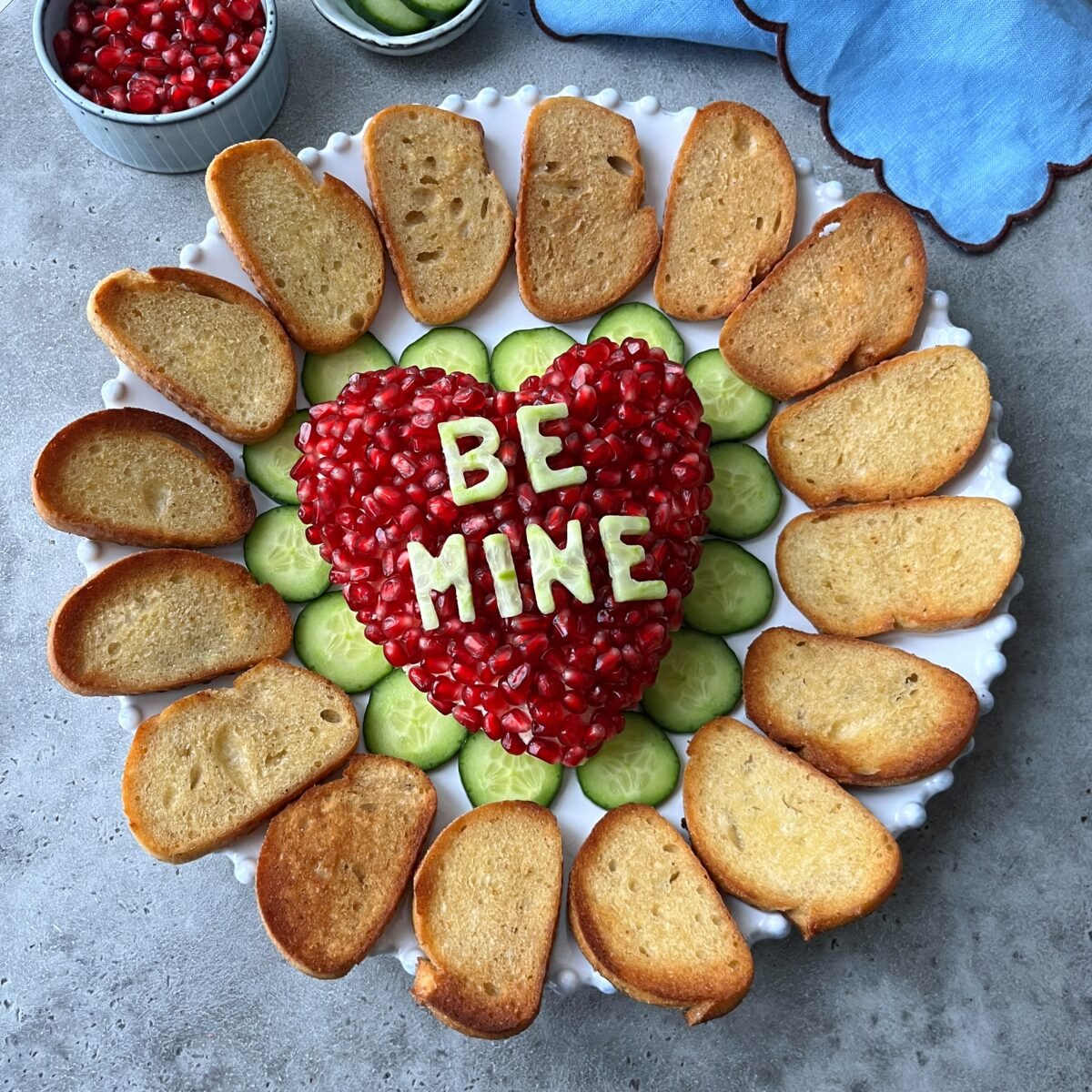 A plate with a heart-shaped cheese ball topped with pomegranate seeds and BE MINE spelt with cucumber, surrounded by sliced cucumbers and toasted bread.