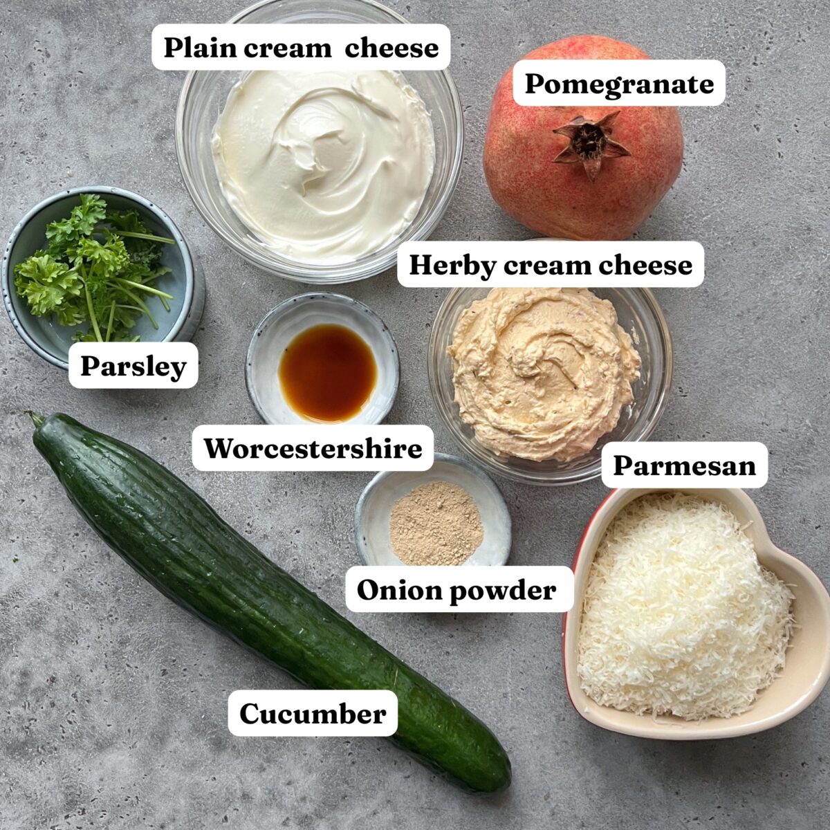 Assorted ingredients on a gray surface including cucumber, parsley, Worcestershire sauce, pomegranate, plain and herby cream cheese, onion powder, and grated parmesan.