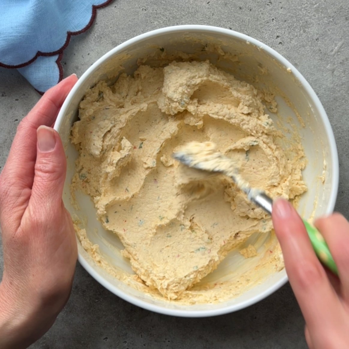 A person mixing a thick, yellow cheese ball in a white bowl with a green-handled spatula on a gray countertop.