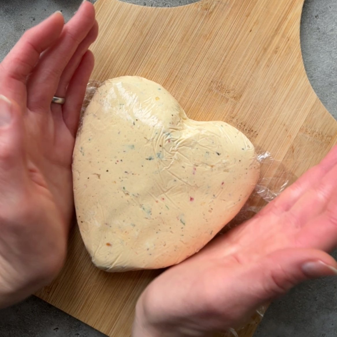 Two hands shape a heart-shaped cheese ball covered in plastic wrap on a wooden cutting board.