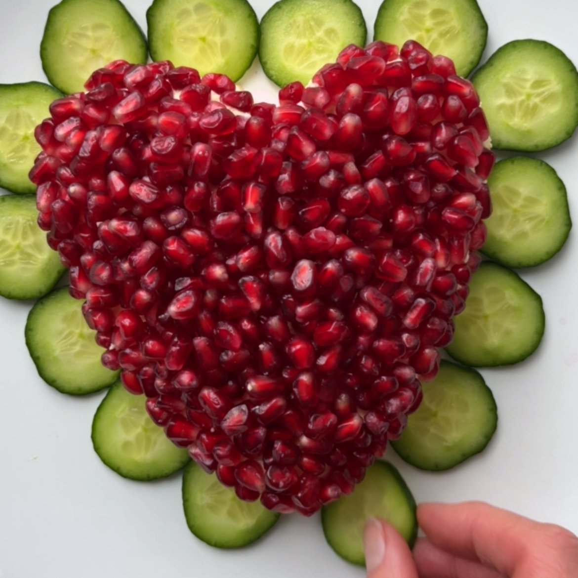A heart shape cheese ball topped with pomegranate seeds is surrounded by cucumber slices on a white surface.