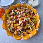 A large round dish filled with a colorful fruit salad, including oranges, apples, kiwi, pomegranate seeds, and blueberries, with orange slices arranged around the edge.
