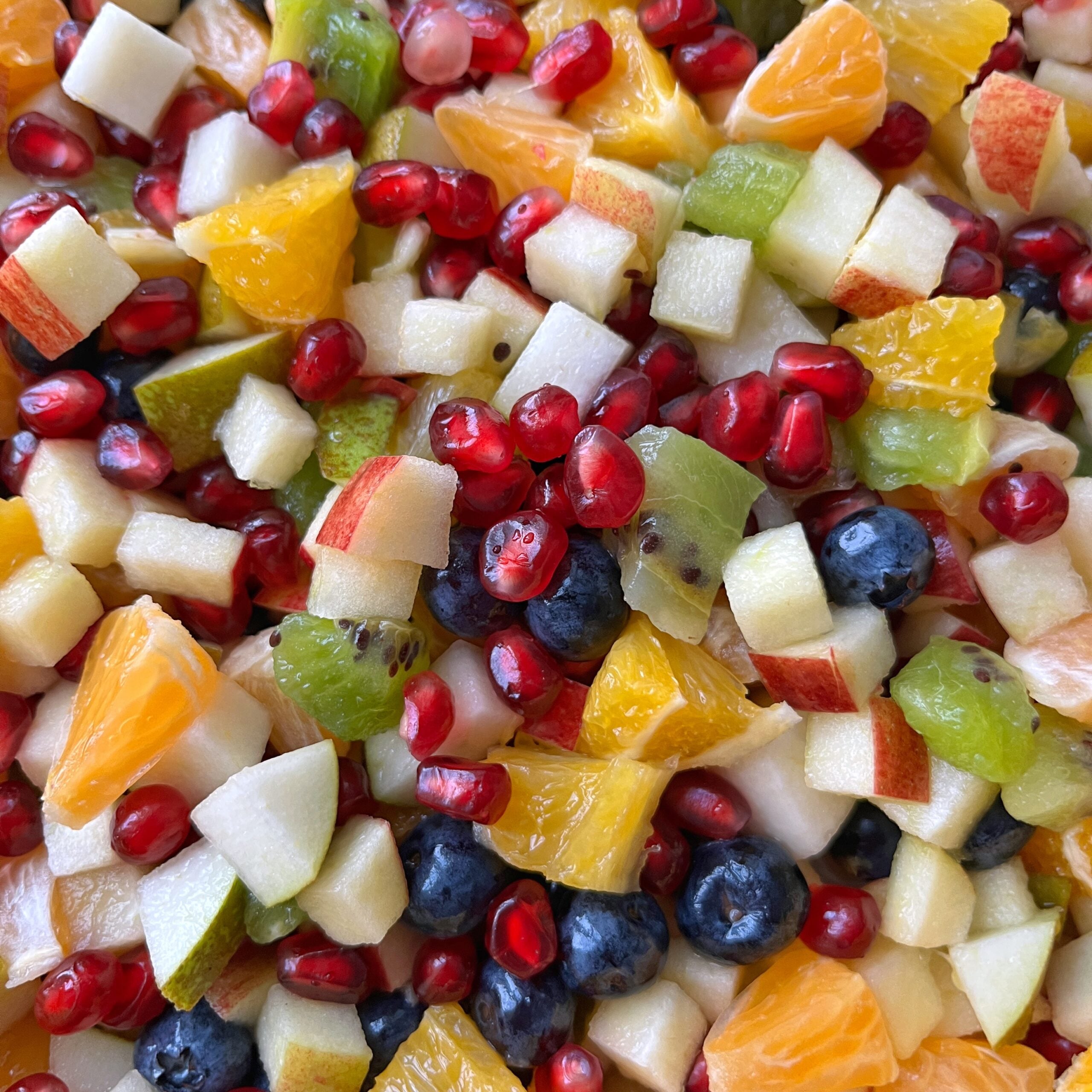 A close-up of a mixed fruit salad with chopped apples, oranges, kiwi, blueberries, pear, and pomegranate seeds.