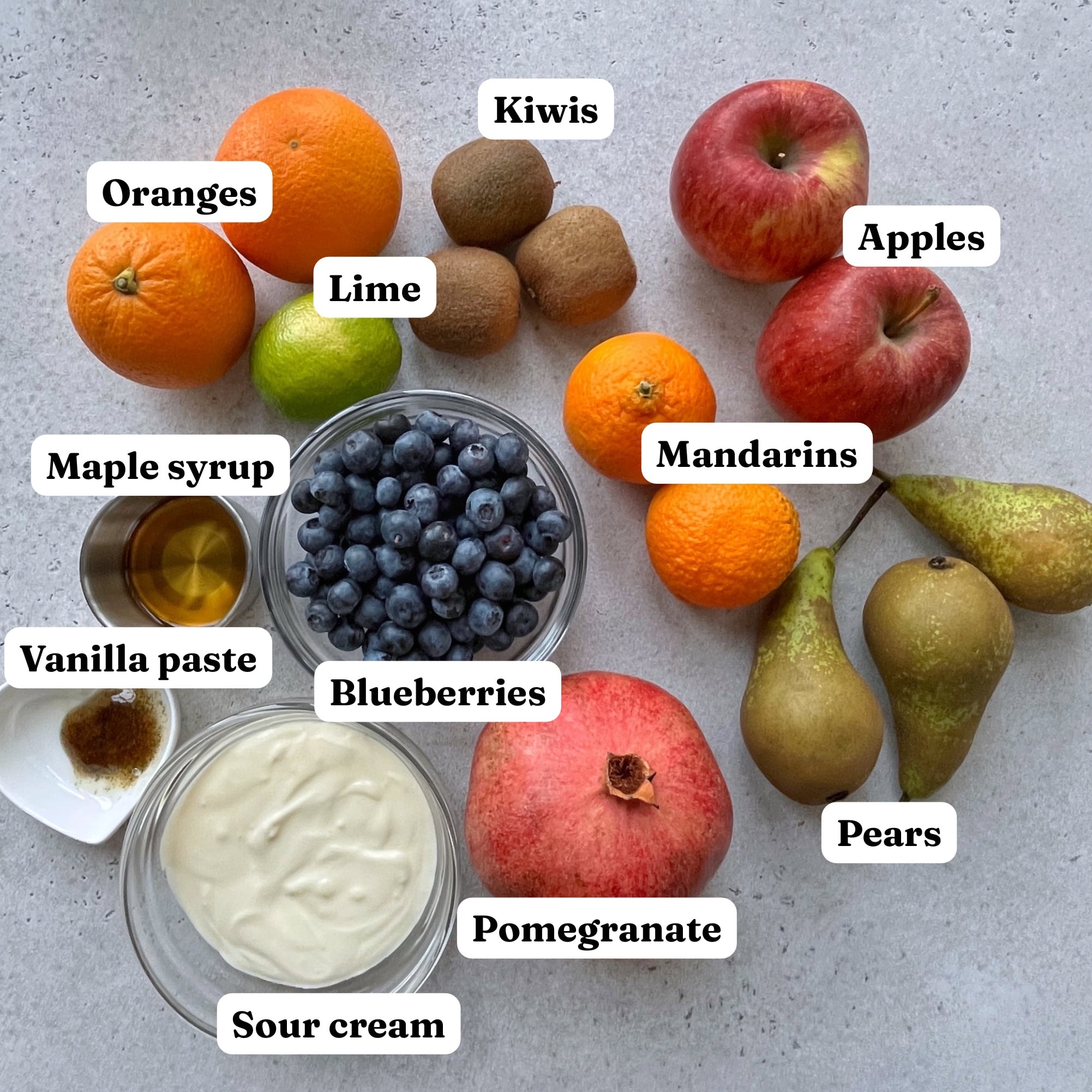 Assorted fruits including oranges, kiwis, apples, lime, mandarins, pears, blueberries, and pomegranate are beautifully arranged for a vibrant Winter Fruit Salad, accompanied by bowls of sour cream, vanilla paste, and maple syrup on a light surface.