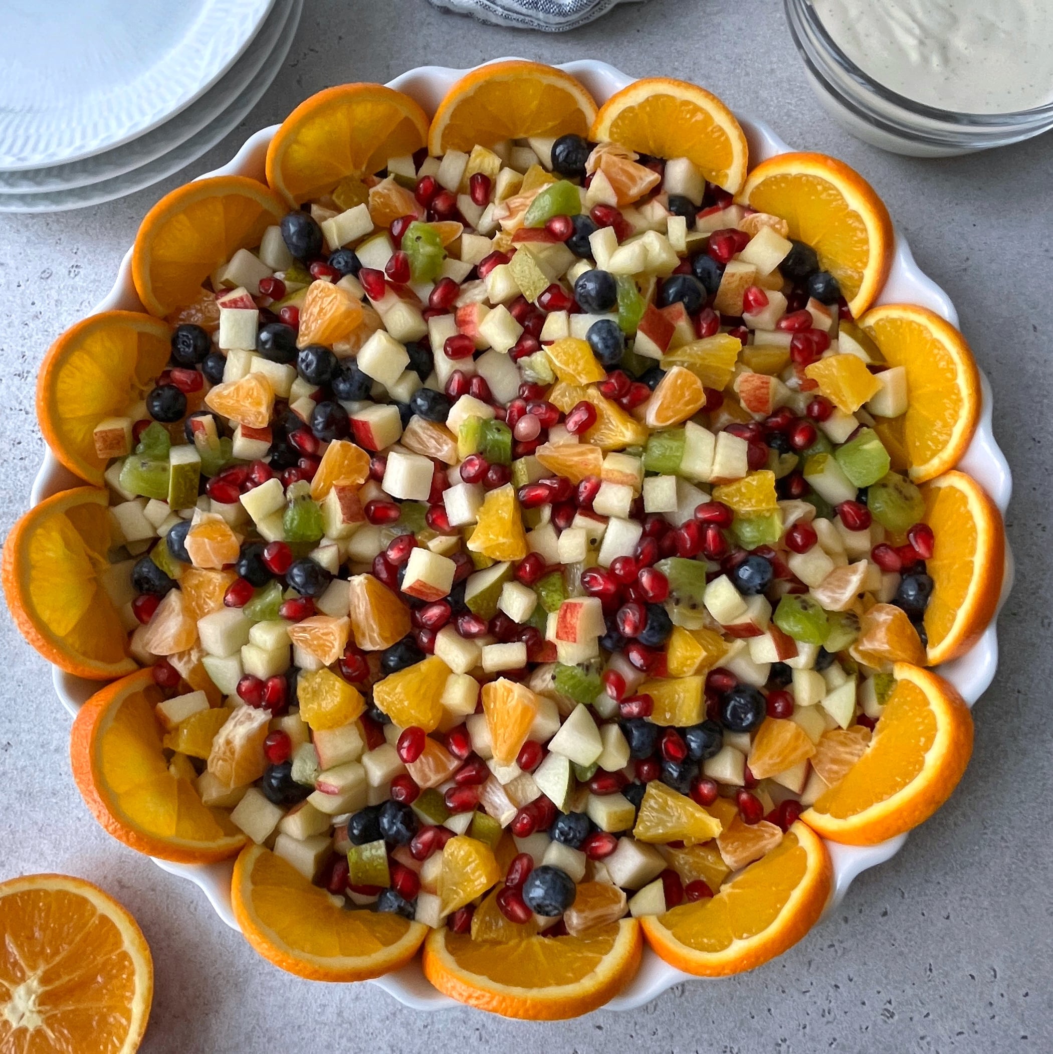A large round dish filled with assorted chopped fruit salad, garnished with orange slices around the edge, sits on a gray surface beside plates and a bowl of dressing.