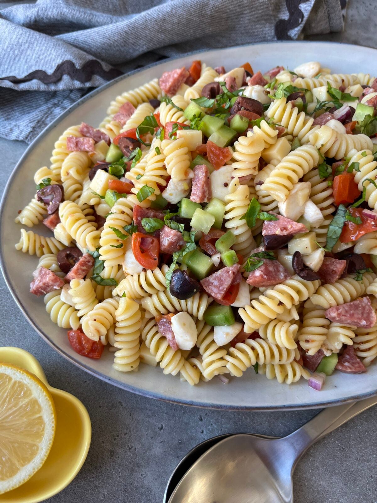 A plate of pasta salad with fusilli, salami, cherry tomatoes, cucumbers, olives, mozzarella, and fresh herbs sits next to a gray napkin, lemon slices, and a serving spoon.