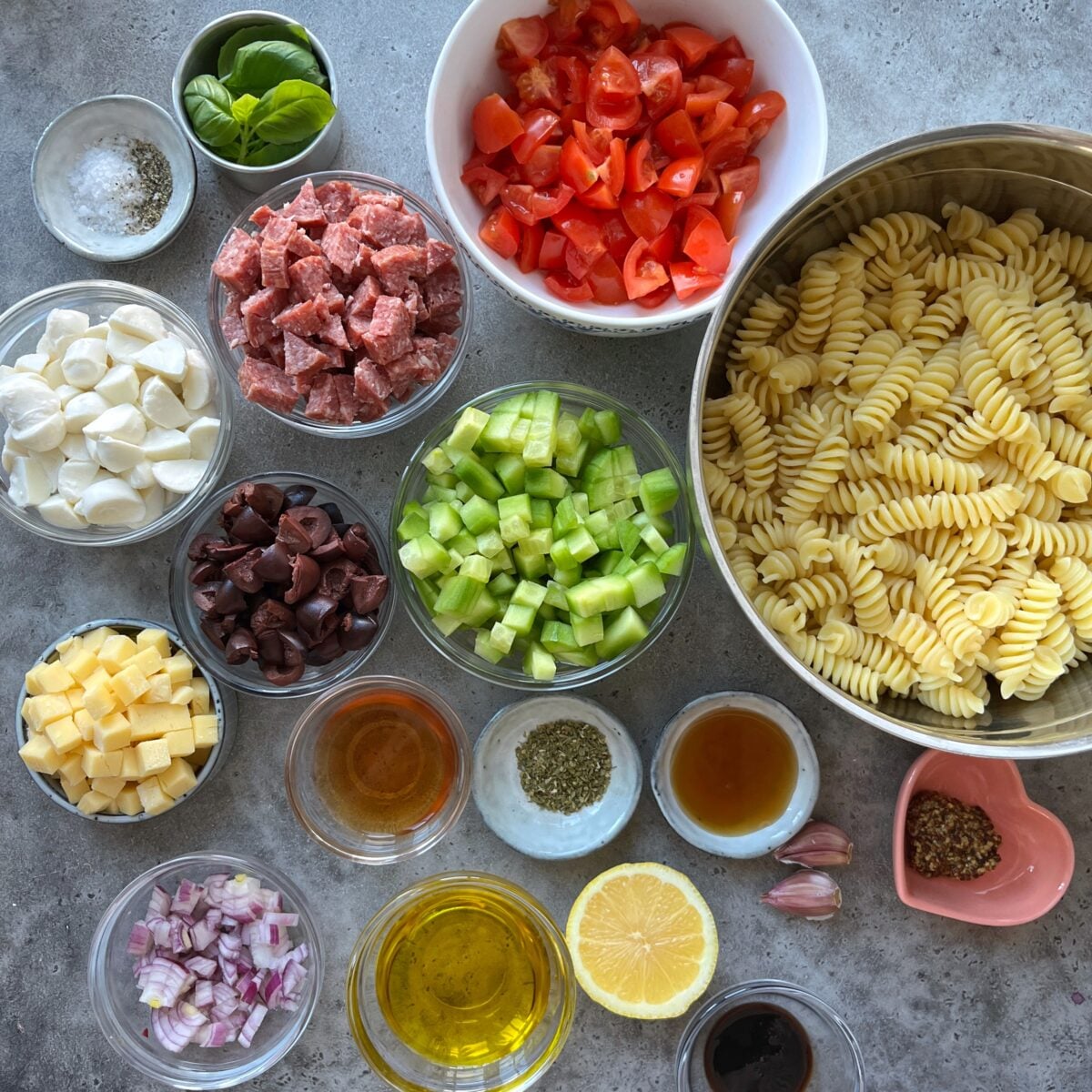 Bowls containing cooked pasta, chopped vegetables, salami, cheese, herbs, olive oil, and seasonings are arranged on a gray surface, ready for a Cold Italian Pasta Salad meal preparation.