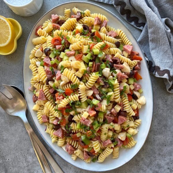 Oval platter with Cold Italian Pasta Salad featuring fusilli mixed with diced meats, cheese, olives, cucumber, tomatoes, set on a gray surface.