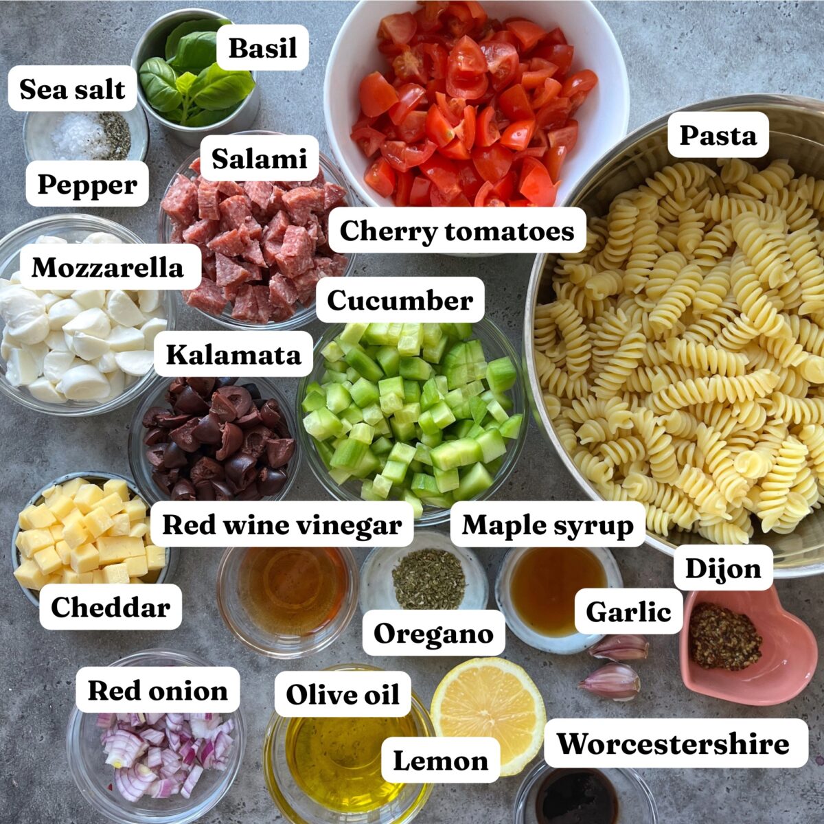 Top view of labeled ingredients for Cold Italian Pasta Salad, including pasta, vegetables, cheeses, meats, herbs, olive oil, and assorted seasonings arranged on a countertop.