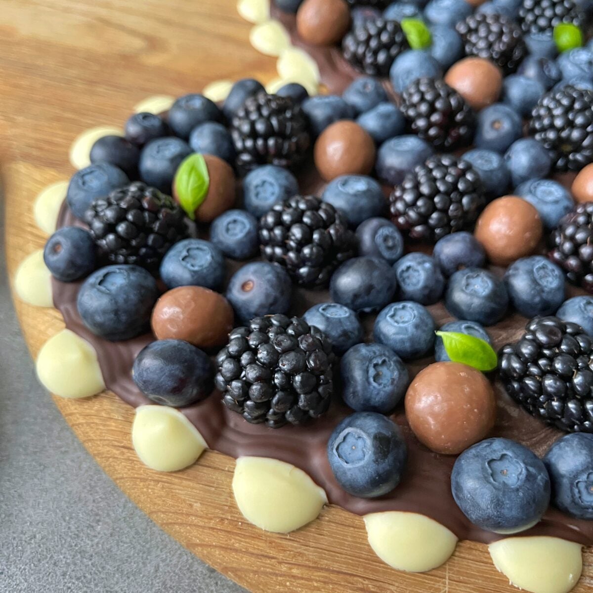 A chocolate bark topped with fresh blueberries, blackberries, round chocolate candies, and small green basil leaves, on a wooden board.