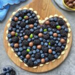 A heart-shaped wooden board topped with blueberries, blackberries, basil leaves, and chocolate candies on a layer of white and dark chocolate.