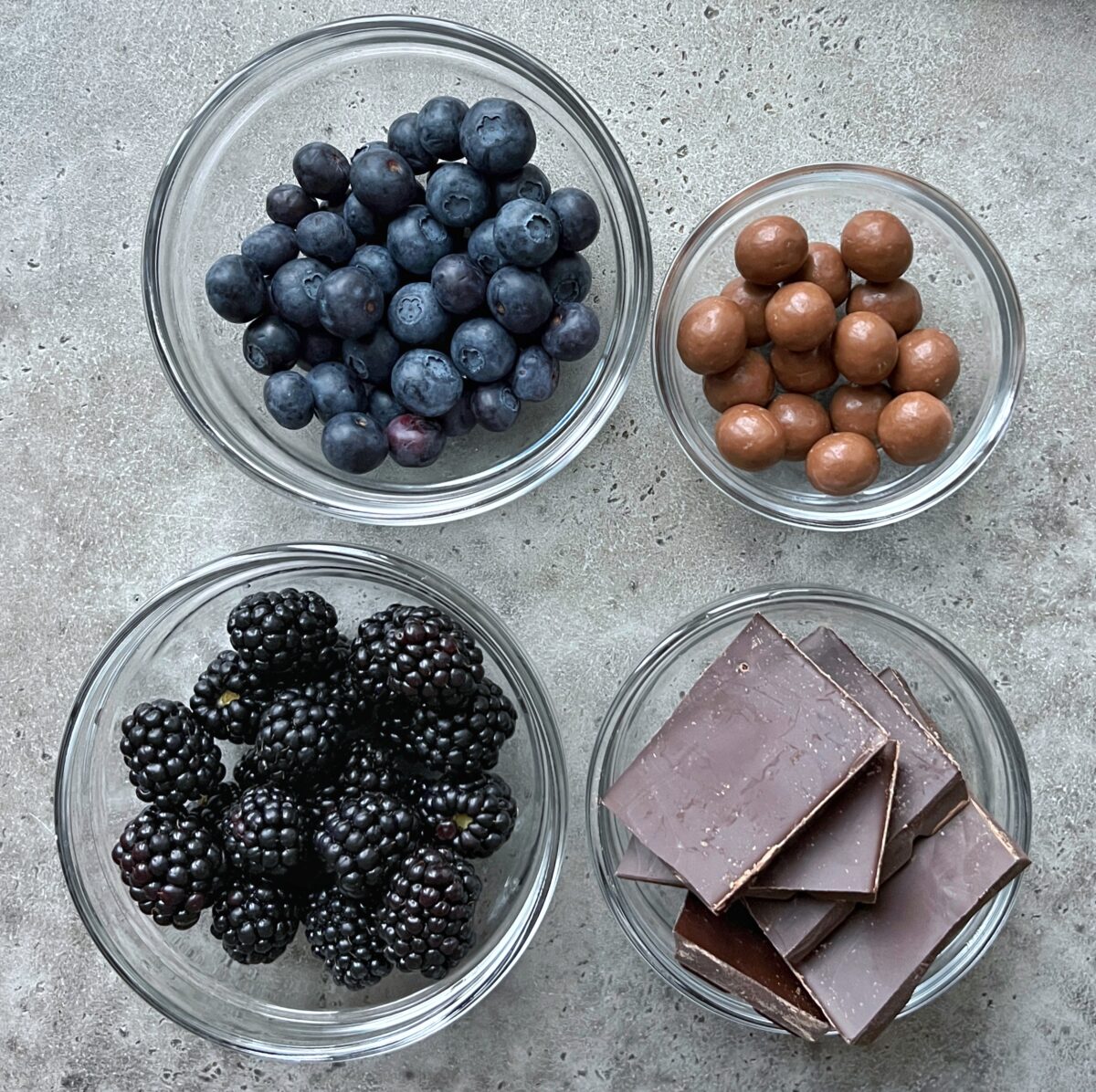 Four glass bowls containing blueberries, chocolate-covered balls, blackberries, and pieces of milk chocolate on a grey surface. 