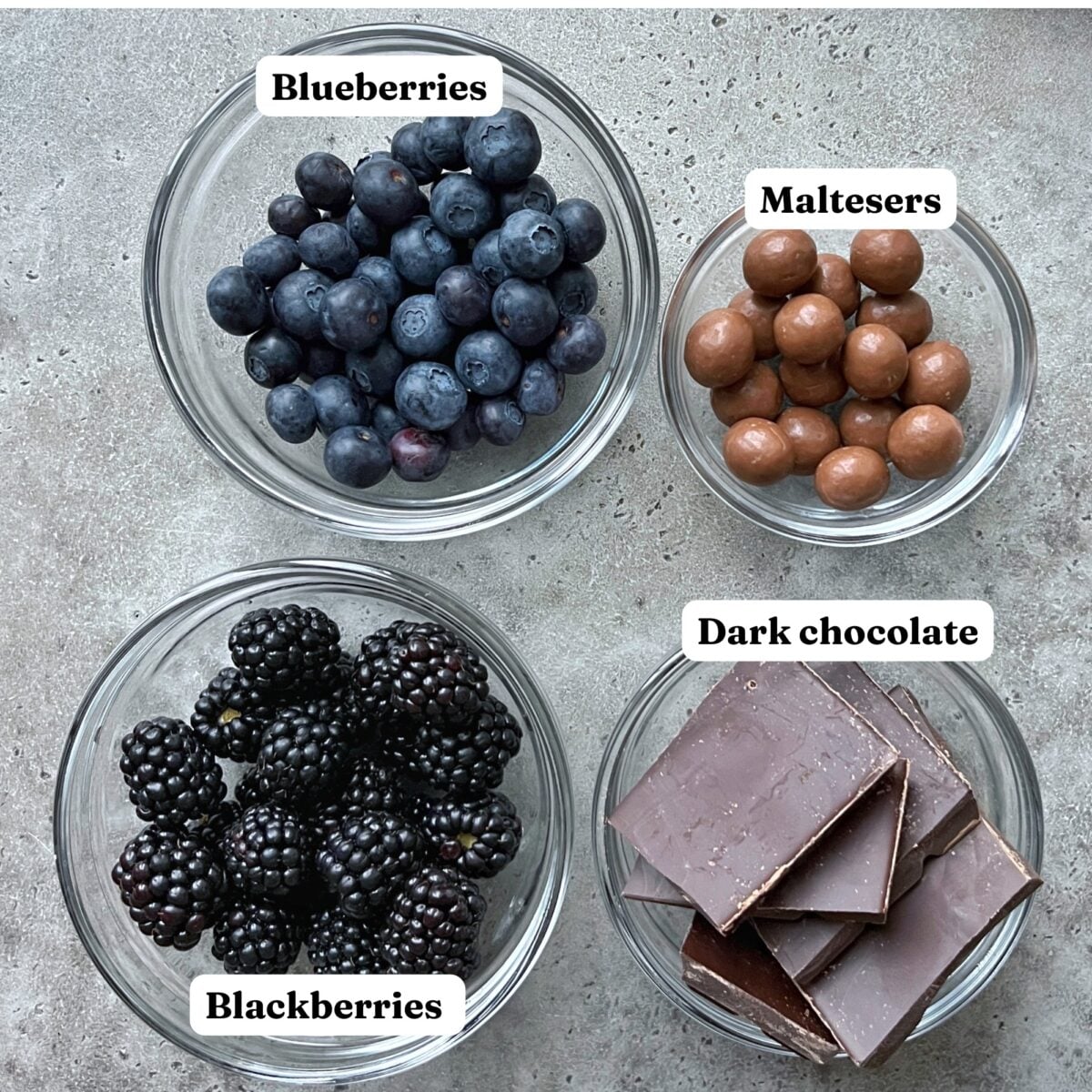 Four glass bowls containing blueberries, Maltesers, blackberries, and pieces of dark chocolate bark are arranged on a grey surface. Each bowl is labeled with its contents.