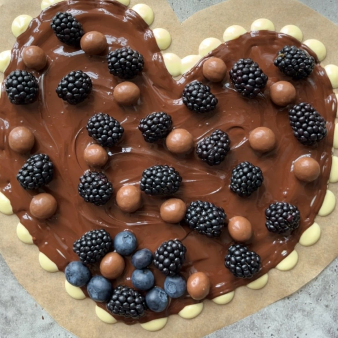 A heart-shaped Dark Chocolate Bark base topped with blackberries, blueberries, and round chocolates, bordered with white chocolate drops.