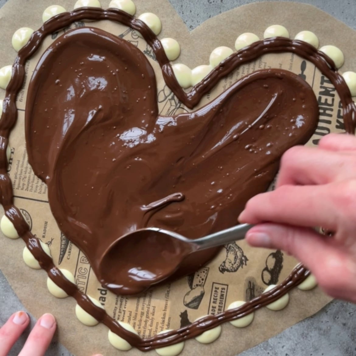 A person spreads melted chocolate inside a heart shape outlined with white and Dark Chocolate Bark dots on parchment paper.