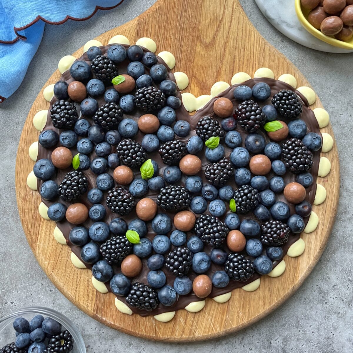 A heart-shaped chocolate bark topped with blueberries, blackberries, and chocolate-covered balls with a white chocolate dot frame.