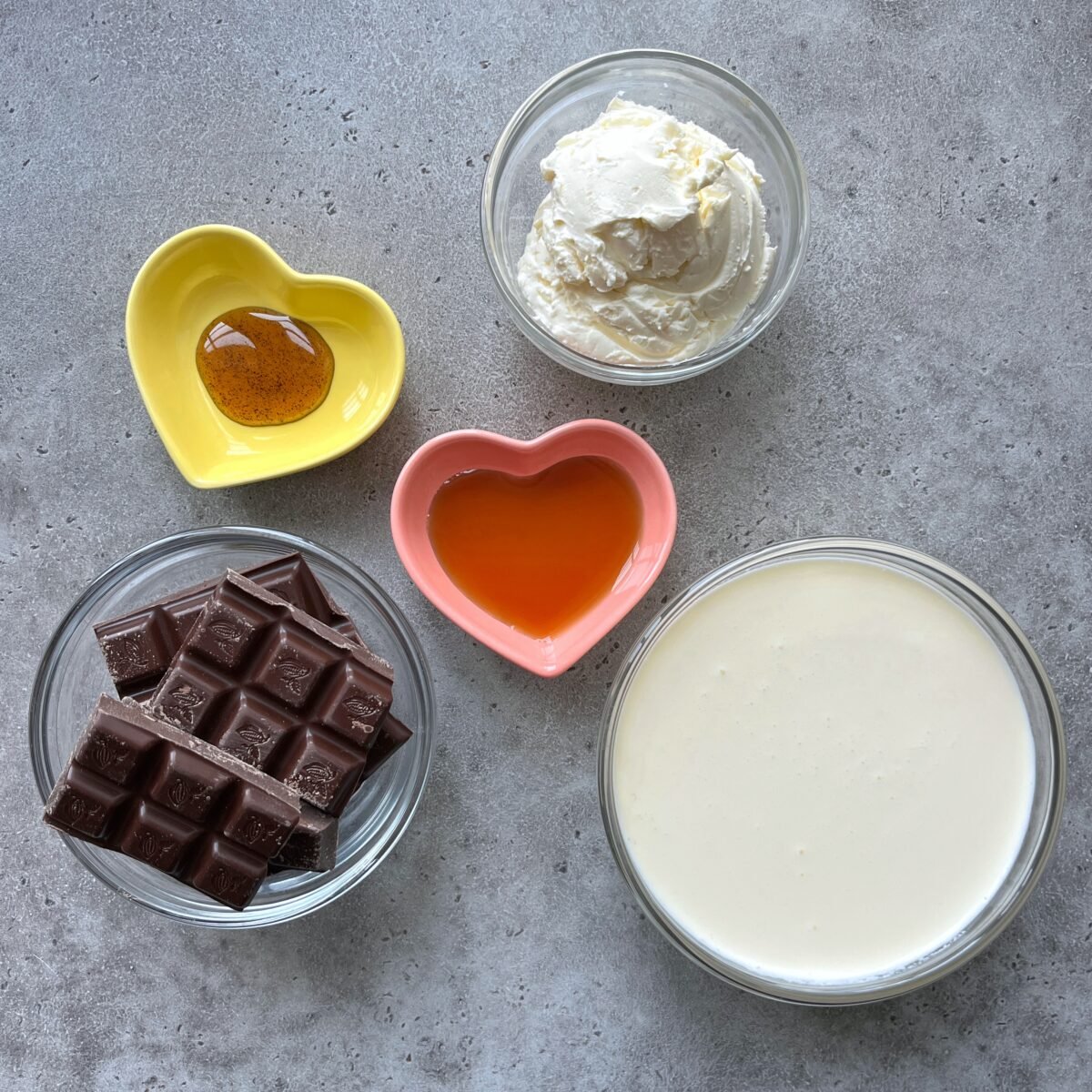 Five bowls containing chocolate pieces, heavy cream, mascarpone, maple syrup, and vanilla paste arranged on a gray surface.