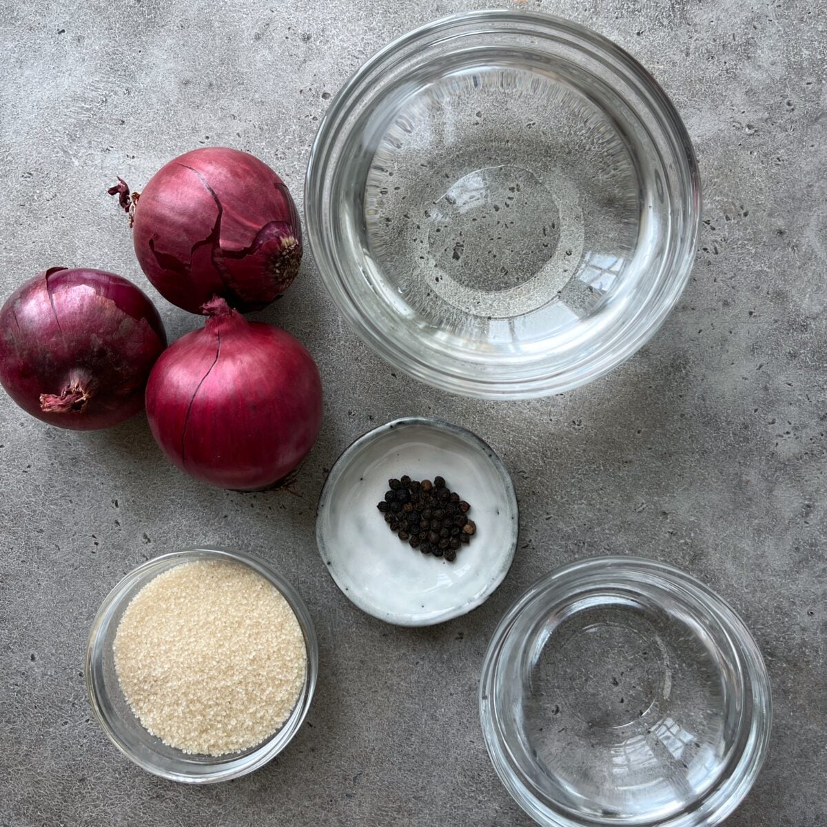 Three red onions, a bowl of water, a small bowl of black peppercorns, a bowl of sugar, and a bowl of vinegar arranged on a gray surface.