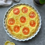 A baked quiche in a white ceramic dish, topped with evenly spaced tomato slices, set on a gray surface.