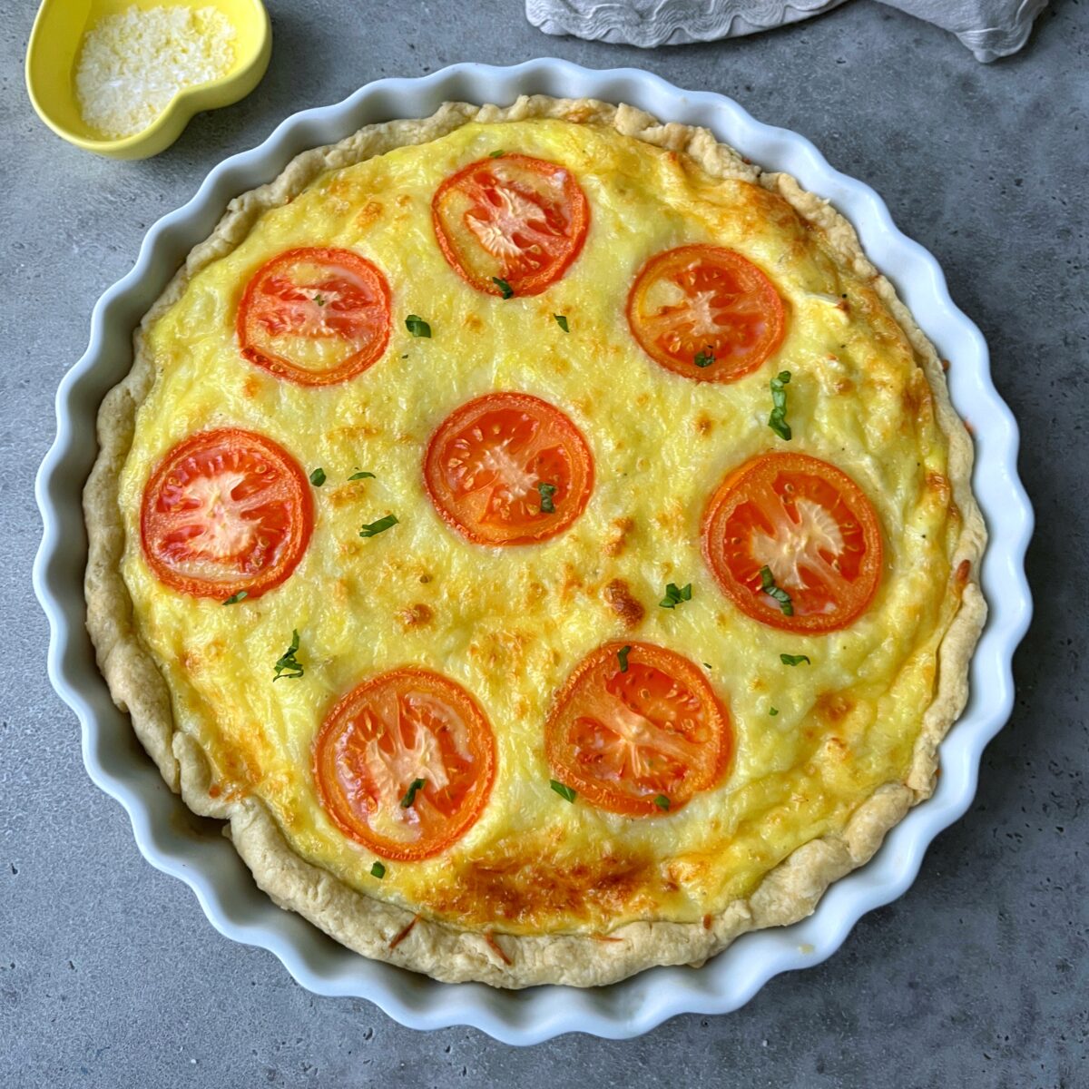 A side view of a baked goat cheese quiche in a white dish topped with sliced tomatoes and chopped herbs.